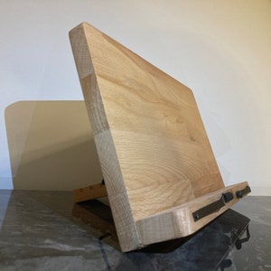 May include: A wooden phone stand with a natural finish and black metal accents. The stand is angled to hold a phone upright.