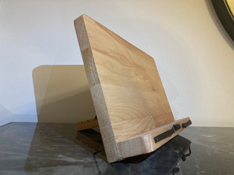 May include: A wooden phone stand with a natural finish and black metal accents. The stand is angled to hold a phone upright.