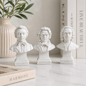 Op de afbeelding: Drie witte bustes van klassieke componisten: Mozart, Beethoven en Bach. Elke buste staat op een rechthoekige voet met de naam van de componist. De bustes worden tentoongesteld op een marmeren oppervlak met een vaas en boeken op de achtergrond.