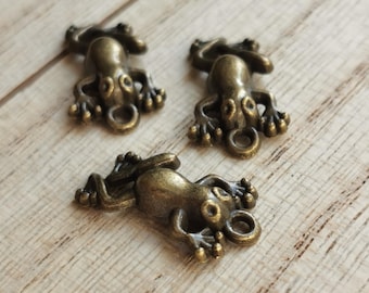 May include: Three bronze-colored frog charms with detailed features, including textured skin and circular loops. The charms are arranged on a light wood-grain surface, showcasing their three-dimensional design.