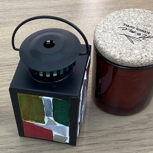May include: A black metal lantern with a mosaic design on the sides, next to a brown glass candle jar with a cork lid. The candle jar has the word "Home" printed on the lid.