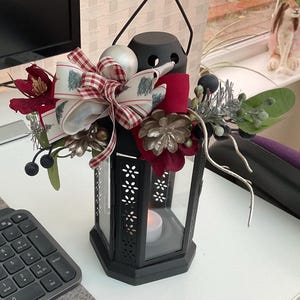 May include: A black metal lantern decorated with a festive floral arrangement. The arrangement features red flowers, silver embellishments, and a bow with a Christmas tree design. A candle is lit inside the lantern.