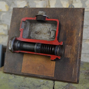May include: A cutaway model of a mechanical device mounted on a wooden board. The device is made of metal and has a red painted exterior. The interior of the device is visible, revealing a spring and other internal components.