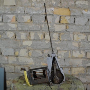 May include: A vintage metal machine with a long, thin rod extending from the top. The machine has a gear and a crank, and is sitting on a green surface.