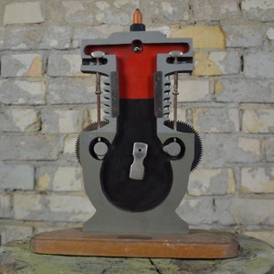 May include: A cutaway model of a four-stroke internal combustion engine. The model is made of metal and painted gray, with red accents. The engine is mounted on a wooden base.
