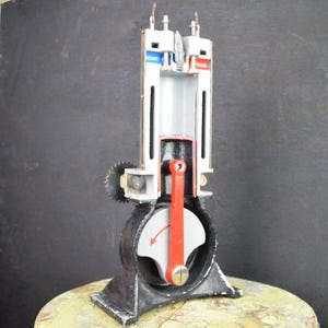 May include: A detailed model of an engine, showcasing internal components. The model is painted in various colors, including red, blue, and silver, to highlight different parts. The base is black, and the model is mounted on a round, textured surface.