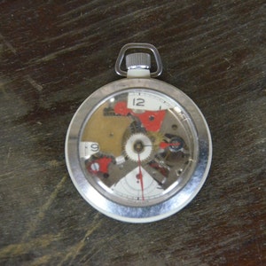 May include: A silver pocket watch with a clear face revealing the inner workings of the watch. The watch face has black numbers and red accents.