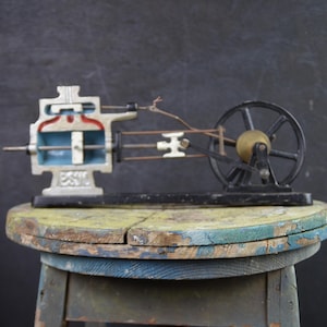 Può includere: Un modello vintage di motore a vapore, con una sezione trasversale dettagliata dei componenti interni. Il modello ha un volano nero, dettagli in ottone e un interno blu e rosso. È posto su uno sgabello di legno usurato.