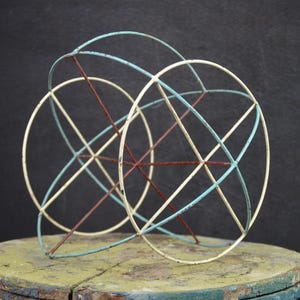 Op de afbeelding: Een vintage metalen sculptuur met elkaar kruisende cirkelvormige vormen. De sculptuur heeft een combinatie van kleuren, waaronder gebroken wit, lichtblauw en roestbruin. Het geometrische ontwerp creëert een interessante visuele weergave, geschikt voor interieurdecoratie.