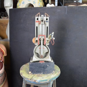Può includere: Un modello di motore sezionato, che mostra i componenti interni. Il modello è principalmente argentato con accenti rossi e arancioni, montato su una base nera. È posto su uno sgabello piccolo, rotondo e multicolore. Lo sfondo è grigio scuro.