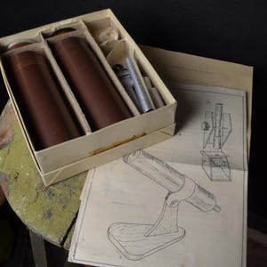 May include: An open box containing two brown cylindrical objects, possibly telescopes, with additional components. The box sits on a weathered, round surface next to a blueprint of a similar device. The items are likely vintage or antique.