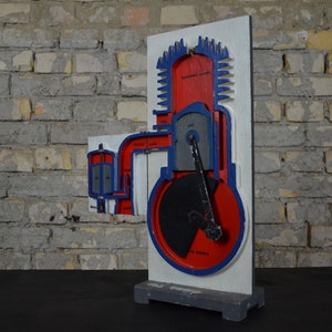 Op de afbeelding: Een vintage houten model van een viertakt-verbrandingsmotor. Het model is geschilderd in rood, blauw en zwart en toont de verschillende fasen van de motorcyclus. Het model is gemonteerd op een houten basis.