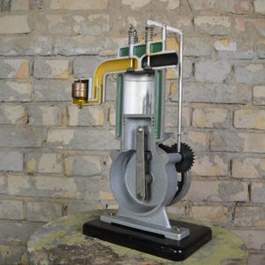 Peut inclure: Modèle de moteur à combustion interne à quatre temps en coupe. Le modèle est en métal et en plastique et montre les différentes parties du moteur, y compris le piston, le vilebrequin et la bielle. Le moteur est monté sur une base noire.