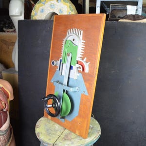 May include: A wooden wall art piece with a mechanical design. The artwork features a brown wooden base with a green, blue, and silver design. A black steering wheel is attached to the front. The piece is mounted on a small, round stool.