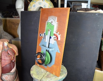 May include: A wooden wall art piece with a mechanical design. The artwork features a brown wooden base with a green, blue, and silver design. A black steering wheel is attached to the front. The piece is mounted on a small, round stool.