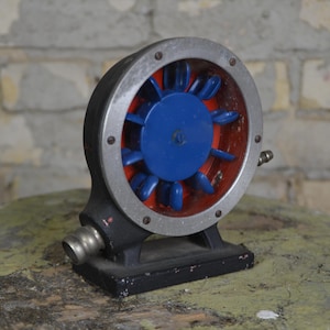 Puede incluir: Un motor eléctrico vintage de color plateado con una hélice de ventilador azul y roja. El motor está montado sobre una base negra.