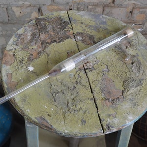 Könnte beinhalten: Ein klares Glasrohr mit einem Korkstopfen auf einer verwitterten Holzoberfläche. Das Rohr ist lang und dünn, mit einer leichten Biegung am Ende. Die Oberfläche ist grün und braun, mit einem Riss, der durch die Mitte verläuft.