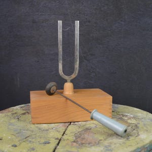 May include: A silver tuning fork with a wooden base and a small mallet. The tuning fork is a U-shaped instrument used to produce a specific pitch. The mallet has a grey handle and a small, round striking head. The items are on a wooden block.