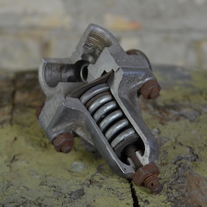 May include: A cutaway view of a metal valve with a spring mechanism. The valve is made of silver metal and has several bolts and nuts. The valve is sitting on a rough, green surface.