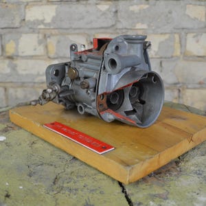 Peut inclure: Un carburateur gris et rouge, coupé en deux, est posé sur un bloc de bois. Le carburateur comporte divers composants métalliques et est présenté contre un mur de briques. Une règle métallique rouge est placée sur le bloc de bois.