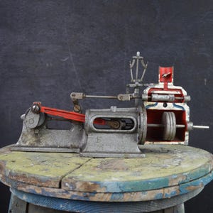 Peut inclure: Modèle de moteur à vapeur vintage, rouge et argenté. Il comprend un volant, un piston et d'autres composants mécaniques. Exposé sur une surface en bois ronde et patinée, sur fond sombre. Un objet de collection.