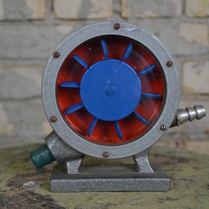 Può includere: Un ventilatore vintage in metallo grigio con un'elica rossa e blu. Il ventilatore ha una base rotonda grigia e un piccolo tubo grigio che si estende dal lato.