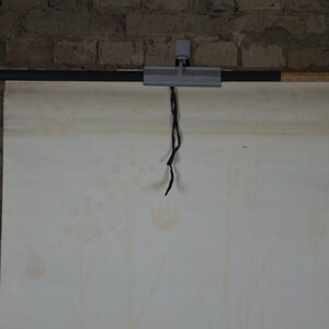 May include: A vintage educational chart, suspended from a metal and wood bar with a black cord. The chart is off-white with age spots, mounted against a brick wall. The bar is held by a metal clamp.