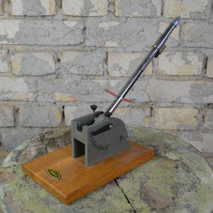 Könnte beinhalten: Ein Vintage-Werkzeug aus Metall und Holz mit silbernem Hebel und grauem Sockel, montiert auf einem Holzsockel. Das Werkzeug hat eine gebogene Skala und steht vor einer verwitterten Backsteinmauer.