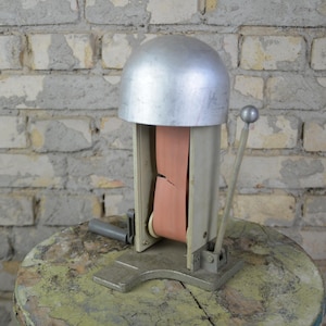 Könnte beinhalten: Eine antike Hutformmaschine aus Metall mit einer silberfarbenen Metallkuppel und einem rosa Stoffband. Die Maschine steht auf einer Holzoberfläche.