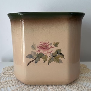 May include: A square ceramic planter featuring a floral design. The planter has a light beige body with a green rim and a painted pink peony with green leaves. The planter is on a white surface.