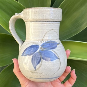 May include: A handmade ceramic jug with a light beige glaze. The jug features a blue floral design and a curved handle. The jug is held in a hand against a background of green leaves. The jug is approximately 15 cm tall.