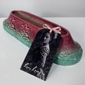 May include: An elongated ceramic planter with a gradient of deep magenta and teal, featuring embossed floral motifs. A rectangular tag with a black and white portrait of a person is attached by a light pink ribbon, displaying the text Love, Laney xo.
