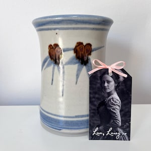 May include: A cylindrical ceramic vase with a cream-coloured glaze, featuring blue horizontal bands at the rim and base, and brown floral motifs. A rectangular black and white photo tag with a pink ribbon displays a portrait and the text Love, Laney xo.