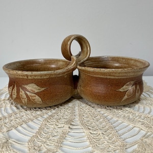 May include: A handcrafted, two-section ceramic serving dish with a handle. The dish is a warm brown colour with a leaf design on each side. The handle is a loop shape, and the dish sits on a cream-coloured, crocheted doily.