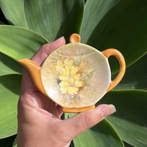 May include: A ceramic teapot-shaped trinket dish in yellow and orange. The dish has a floral design with yellow flowers and green leaves. The handle and spout are orange. The dish is held in a hand against a green background.