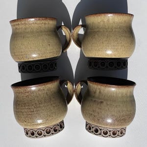 May include: A set of four ceramic mugs with a rounded body and a flared rim. The upper portion features a speckled tan and brown glaze, while the base is a dark brown with a repeating embossed circular pattern around the circumference of each mug.