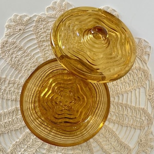 May include: A decorative amber glass dish with a lid. The dish and lid feature a textured, swirling pattern. The round dish has a star-shaped design in the center. The lid has a small knob. The dish is on a cream crocheted doily.