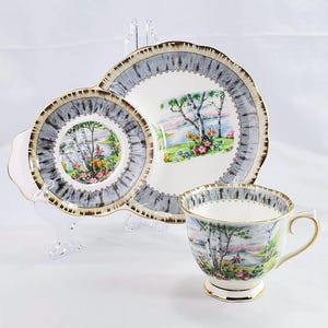 Puede incluir: Juego de taza y platillo vintage con dise&ntilde;o de paisaje. El juego incluye una taza, un plato de dos partes y un platillo. Los bordes est&aacute;n adornados con dorado. El paisaje presenta &aacute;rboles, agua y flores, con un borde gris y blanco.