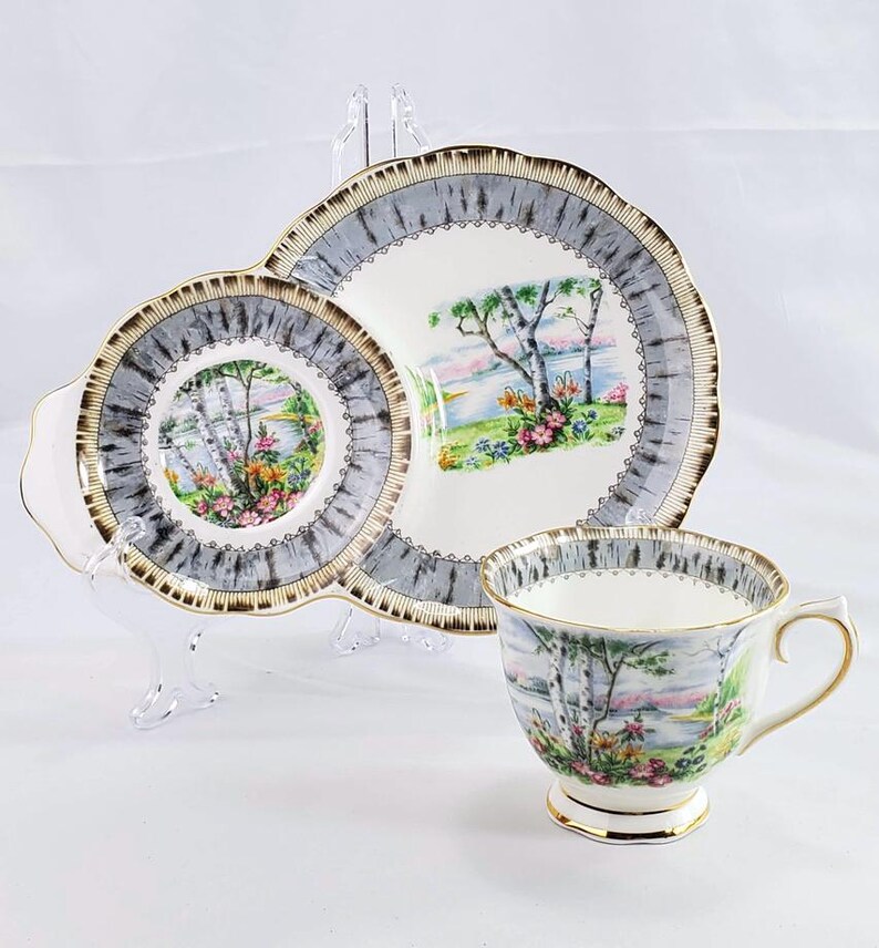 Puede incluir: Juego de taza y platillo vintage con dise&ntilde;o de paisaje. El juego incluye una taza, un plato de dos partes y un platillo. Los bordes est&aacute;n adornados con dorado. El paisaje presenta &aacute;rboles, agua y flores, con un borde gris y blanco.