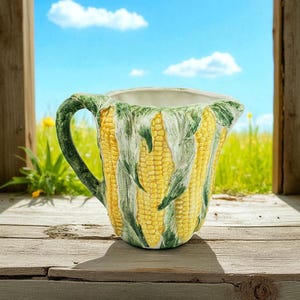 Könnte beinhalten: Eine Keramikkrug mit einem Maiskolben-Design. Der Krug ist gelb mit grünen Blättern und einem grünen Griff. Der Hintergrund zeigt einen blauen Himmel und ein grünes Feld.