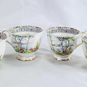 Puede incluir: Cuatro tazas de t&eacute; blancas con un dise&ntilde;o de paisaje con &aacute;rboles, un lago y flores. Las tazas tienen ribetes dorados en el borde, el asa y la base. Las tazas de t&eacute; est&aacute;n dispuestas en fila sobre una superficie blanca.