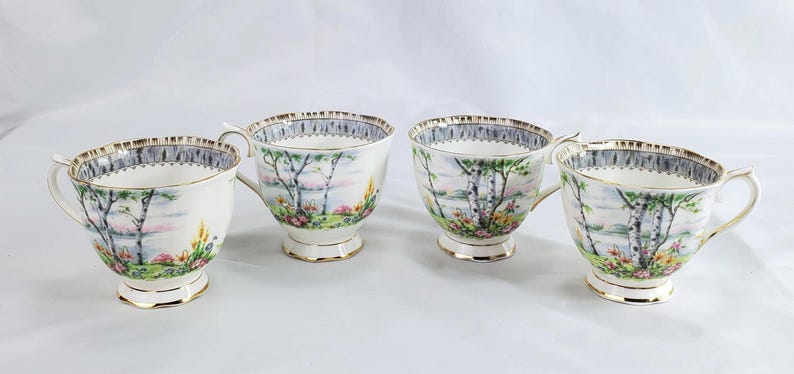 Puede incluir: Cuatro tazas de t&eacute; blancas con un dise&ntilde;o de paisaje con &aacute;rboles, un lago y flores. Las tazas tienen ribetes dorados en el borde, el asa y la base. Las tazas de t&eacute; est&aacute;n dispuestas en fila sobre una superficie blanca.