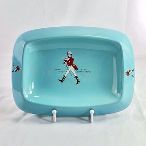 May include: A rectangular, light blue ceramic tray with rounded corners. The tray features a central illustration of a man in a red coat and top hat, with the text "BORN 1820 - STILL GOING STRONG." Small illustrations of the man are on the sides.
