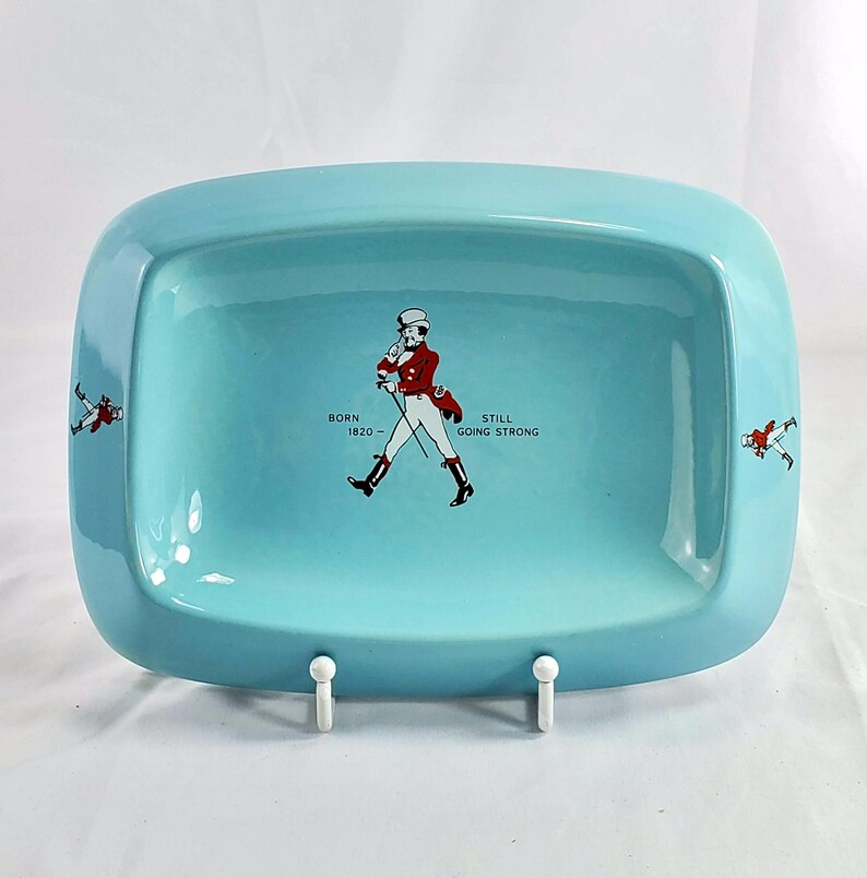 May include: A rectangular, light blue ceramic tray with rounded corners. The tray features a central illustration of a man in a red coat and top hat, with the text "BORN 1820 - STILL GOING STRONG." Small illustrations of the man are on the sides.