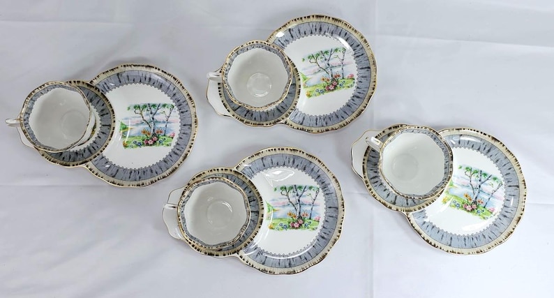 Puede incluir: Cuatro juegos de tazas y platillos. Cada juego incluye una taza blanca con borde dorado, un platillo a juego y un plato peque&ntilde;o. Los platos y platillos tienen un borde azul y gris y una escena de paisaje en el centro.