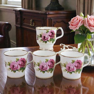Peut inclure: Ensemble de tasses en porcelaine blanche ornées de roses roses et violettes, avec des bordures et des anses dorées. Une grande tasse et un vase de roses roses complètent l'ensemble.