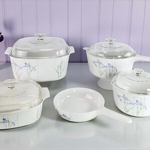 May include: A set of white ceramic cookware with clear glass lids, adorned with a subtle blue floral pattern. The collection includes various pots and pans, ideal for cooking. A vase of purple irises adds a touch of colour.