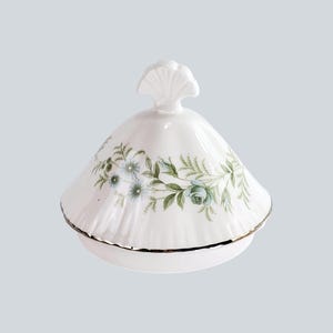 May include: A white porcelain lidded dish with a floral pattern of blue roses and green foliage. The lid has a shell-shaped handle and a gold rim. The dish is likely vintage and suitable for serving or display.