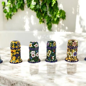 May include: Six small decorative containers with floral designs. The containers are made of metal and have a blue, yellow, green, and gold enamel finish. The containers are arranged in a row on a white marble surface.