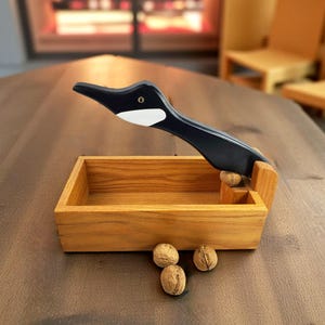 Peut inclure: Un casse-noix en forme d'oie, peint en noir avec des accents blancs, est posé au-dessus d'une boîte en bois. Trois noix sont éparpillées devant la boîte. Le casse-noix est conçu pour casser les noix.